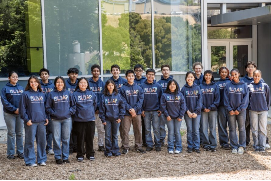 Group shot of many persons all wearing blue sweatshirts reading "BLDAP" and standing in front of glass building.